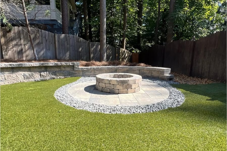 Stone fire pit on a gravel-and-paver circle in a fenced backyard with a low block wall behind it