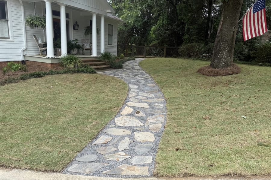 Curved flagstone walkway with a dark paver border crossing a lawn to a white farmhouse porch