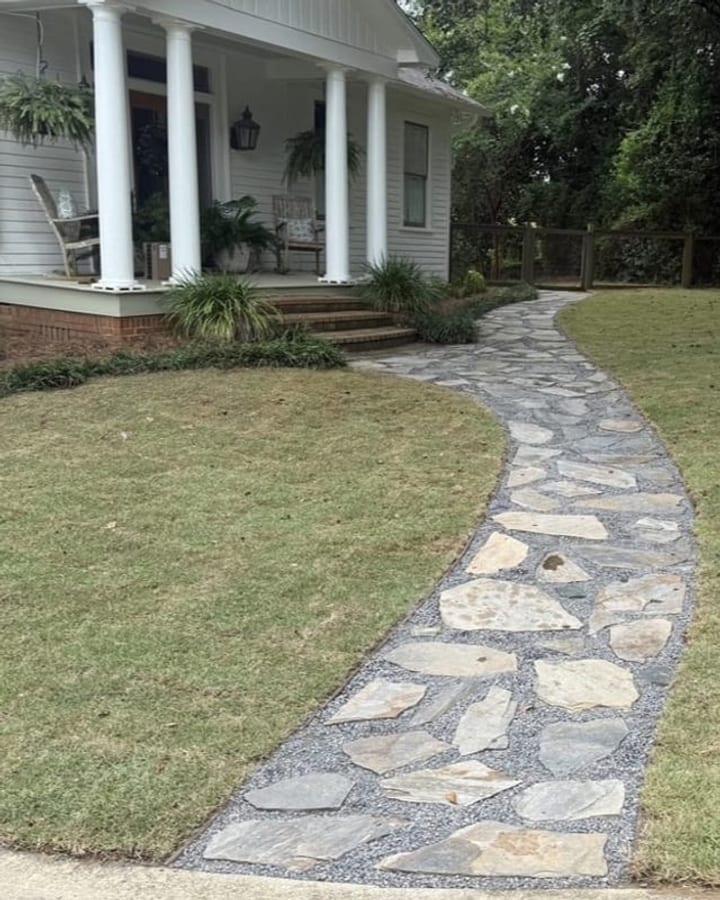 Curved flagstone walkway with a dark paver border crossing a lawn to a white farmhouse porch